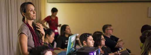 A woman presenting to an audience at the Native American and Indigenous Studies Association using a laptop, with listeners seated at a table focusing on her.